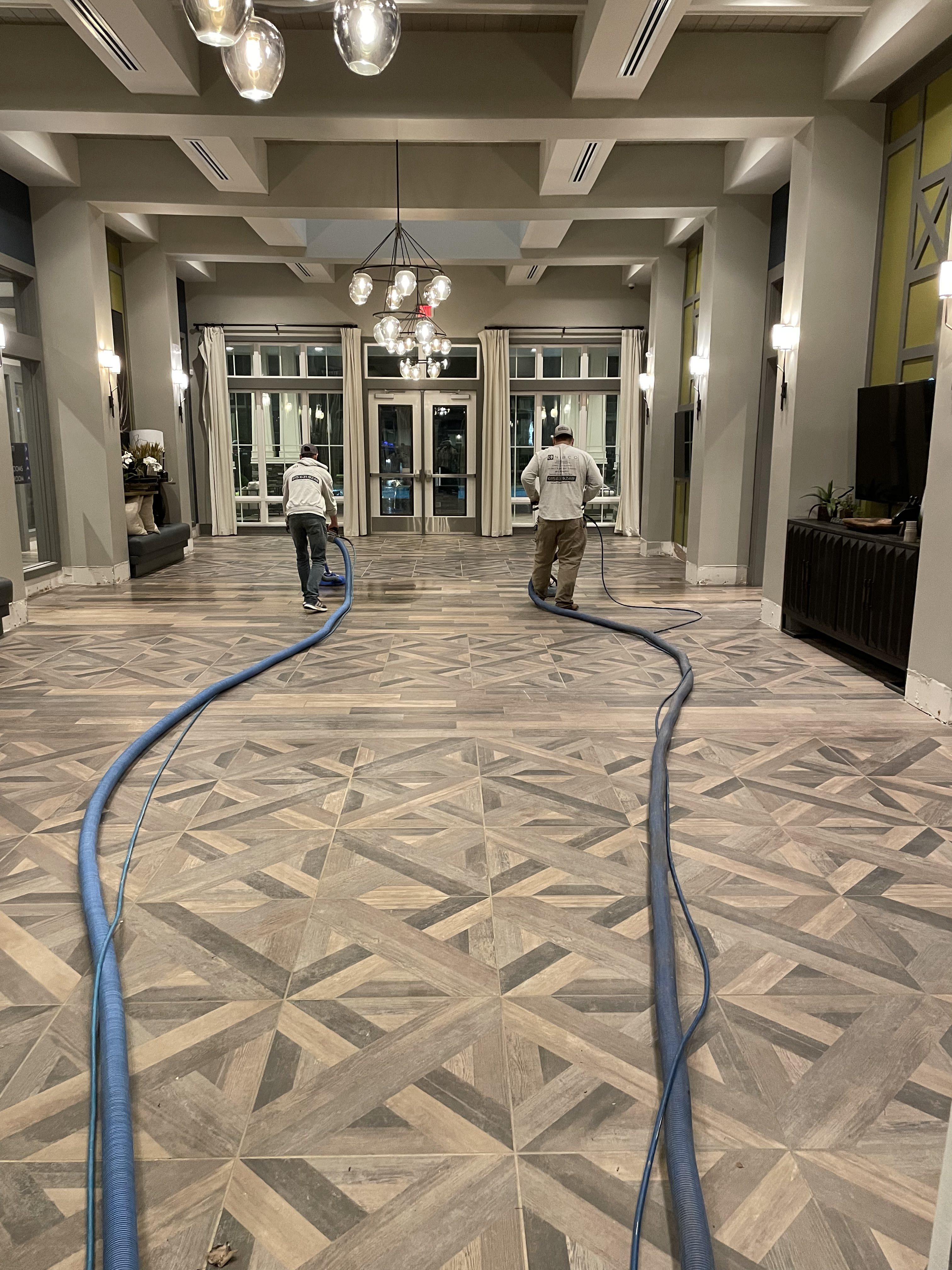 Two people cleaning a tiled floor with hoses in a well-lit lobby with hanging lights and a seating area.